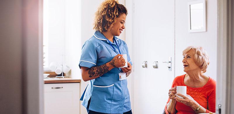 carer with a cup of tea chating with sitting older lady in Kitchen
